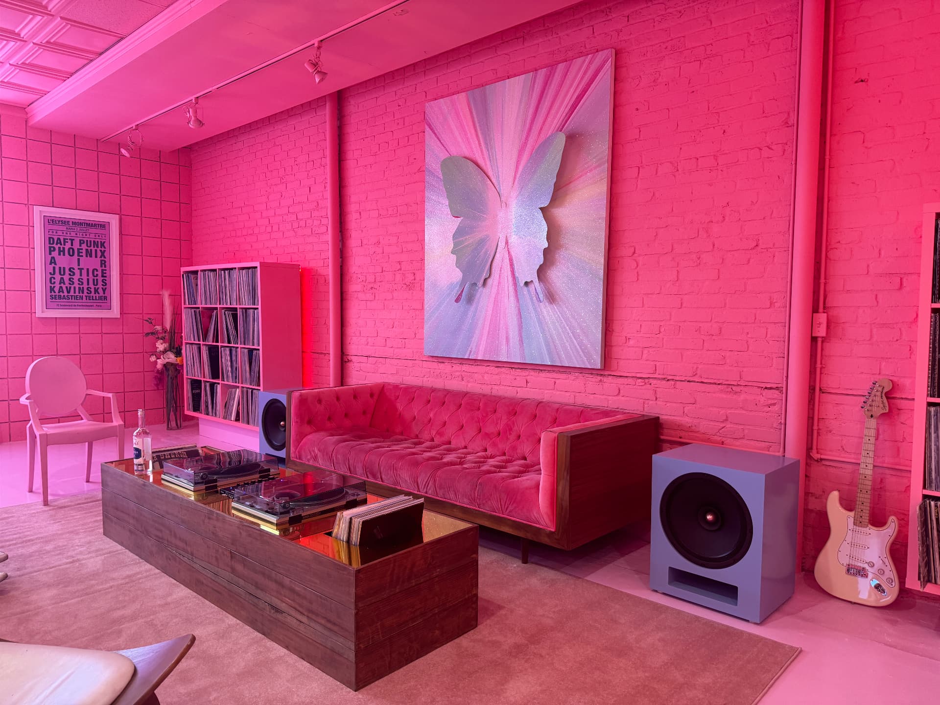 A pair of light blue speakers beside a pink velvet sofa in a pink-lit listening room filled with records